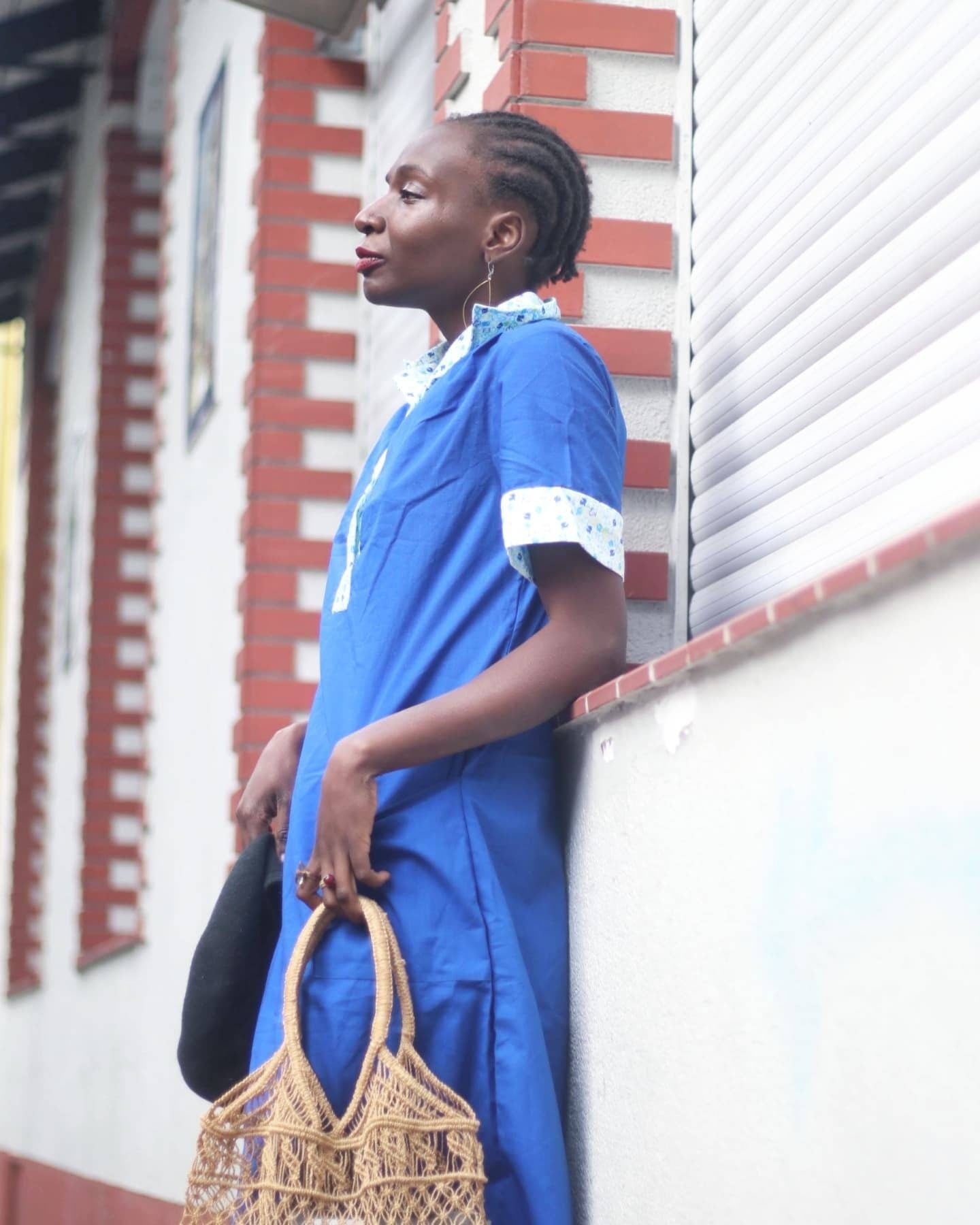 Woman of african heritage in a blue vintage summer dress with printed collar leaning against a brick wall. She holds a woven brown bag in one hand and a black beret in another and looks to the side