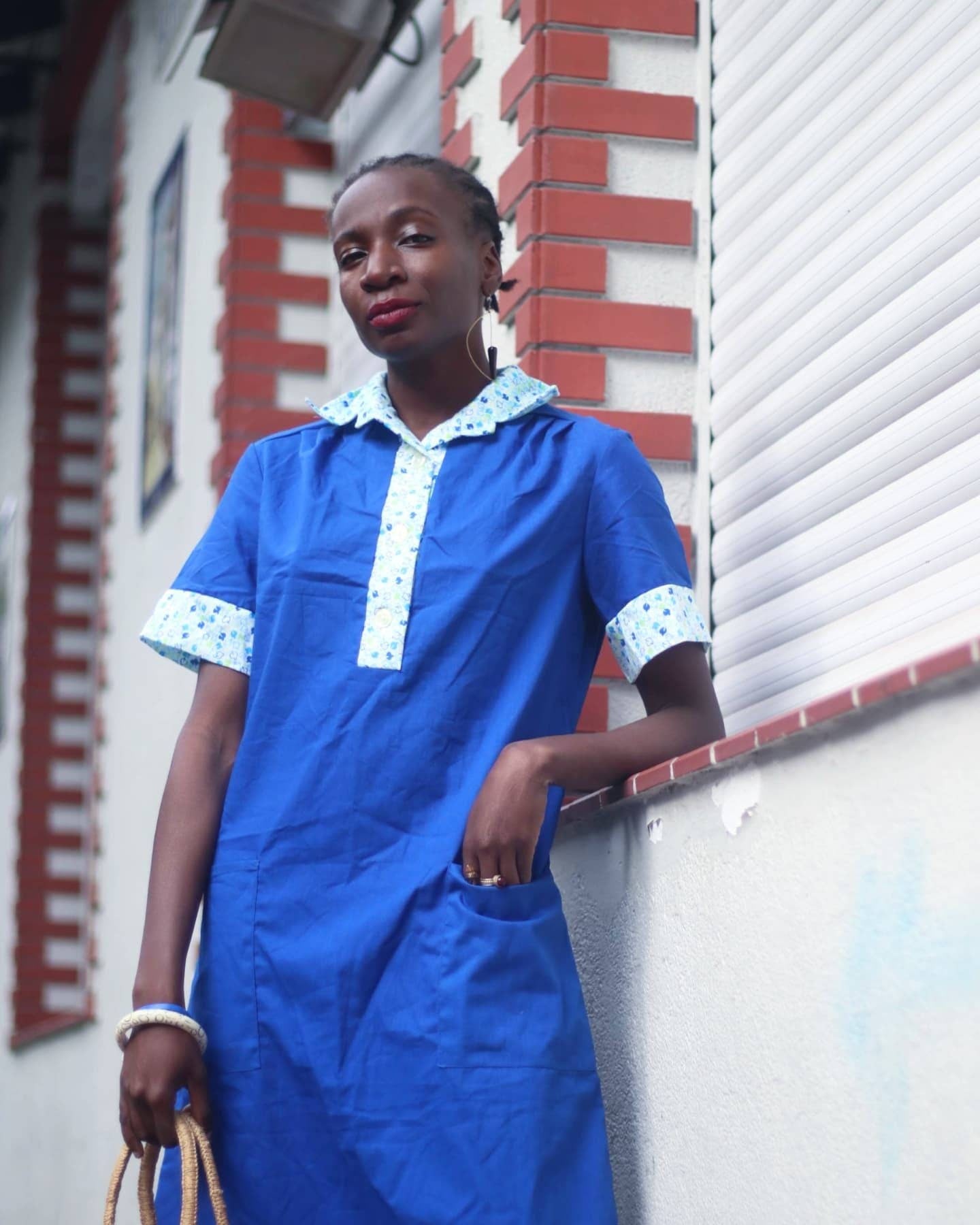 Woman of african heritage in a blue vintage summer dress with printed collar leaning against a brick wall. She holds a woven brown bag in one hand