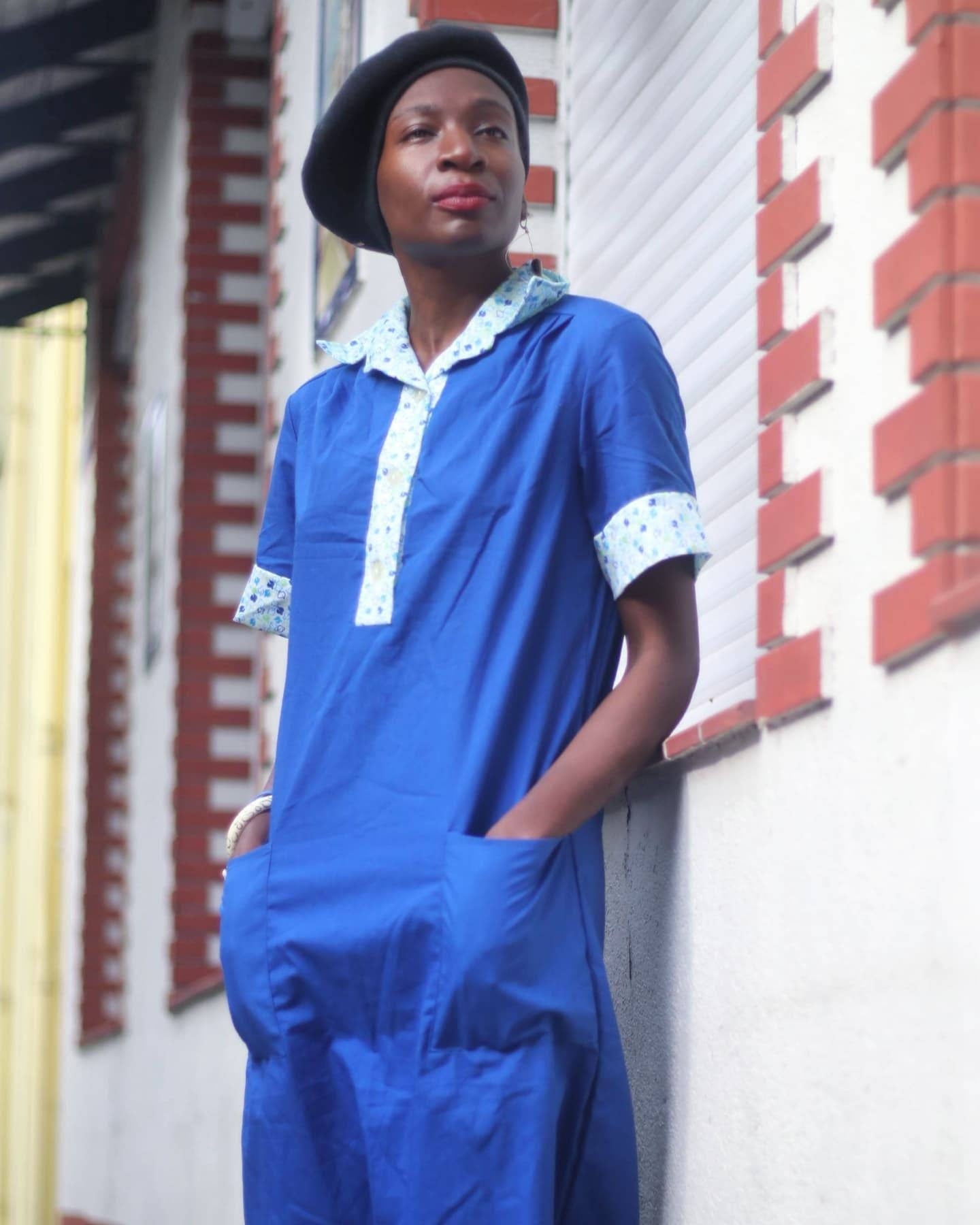 Woman of african heritage in a blue vintage summer dress with printed collar leaning against a brick wall. She is also wearing a black beret and has her hands in the two front pockets of the dress