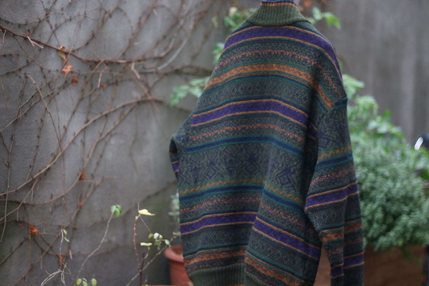 Back view of cozy multi-colored cardigan with green, orange, and purple stripes hanging on a hanger outdoors against a concrete wall with climbing vines. The long-sleeved vintage design can be worn as a coat it is perfect for fall and winter fashion