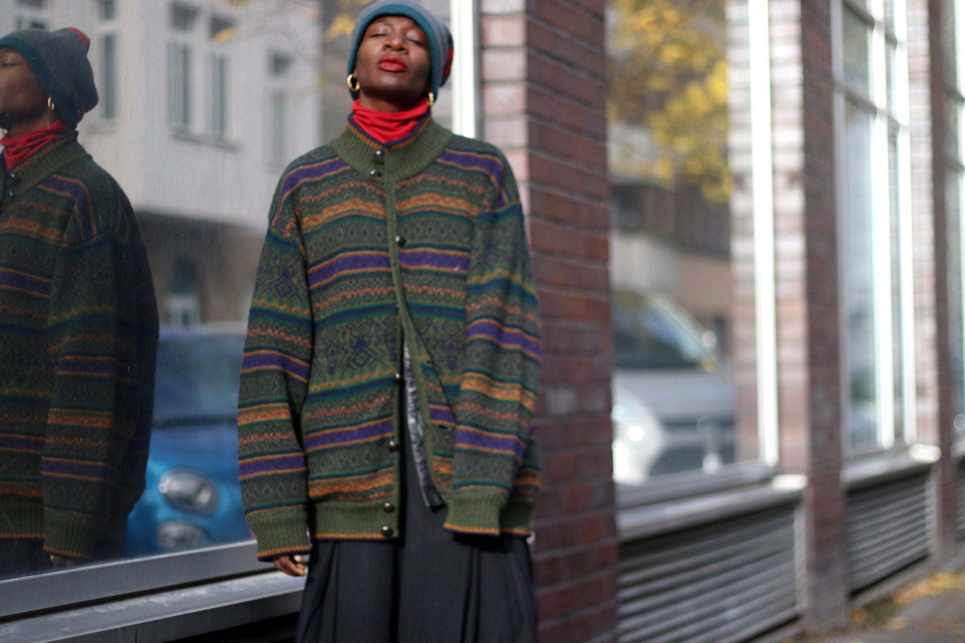 Woman of African Heritage in a vintage patterned green sweater and gray beanie stands casually on an urban street, reflection in window, fall leaves on sidewalk. The hipster long sleeved sweater can be worn as a winter or autumn jacket.