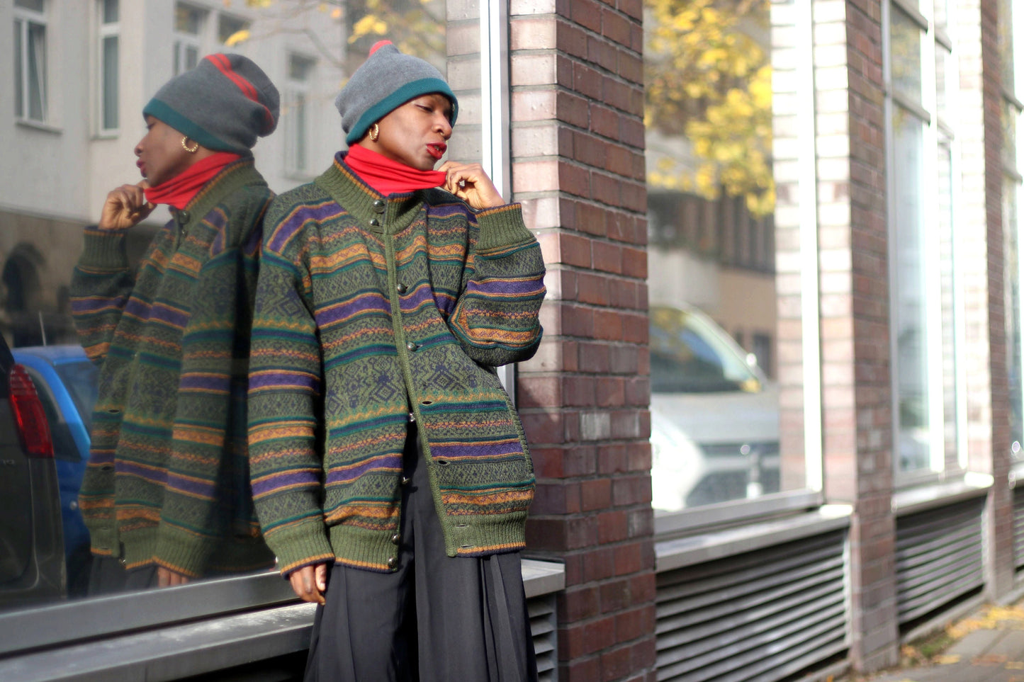 Woman of African Heritage in a vintage patterned green sweater and gray beanie stands casually on an urban street, reflection in window, fall leaves on sidewalk. The hipster long sleeved sweater can be worn as a winter or autumn jacket.