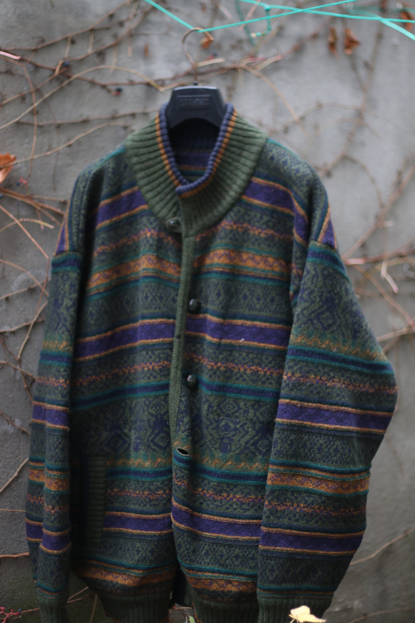 Cozy multi-colored cardigan with green, orange, and purple stripes hanging on a hanger outdoors against a concrete wall with climbing vines. The long-sleeved, vintage design can be worn as a coat and it is perfect for fall and winter fashion.