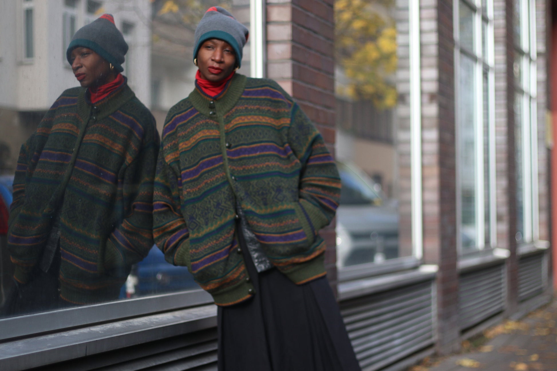 Woman of African Heritage in a vintage patterned green sweater and gray beanie stands casually on an urban street, reflection in window, fall leaves on sidewalk. The hipster long sleeved sweater can be worn as a winter or autumn jacket.