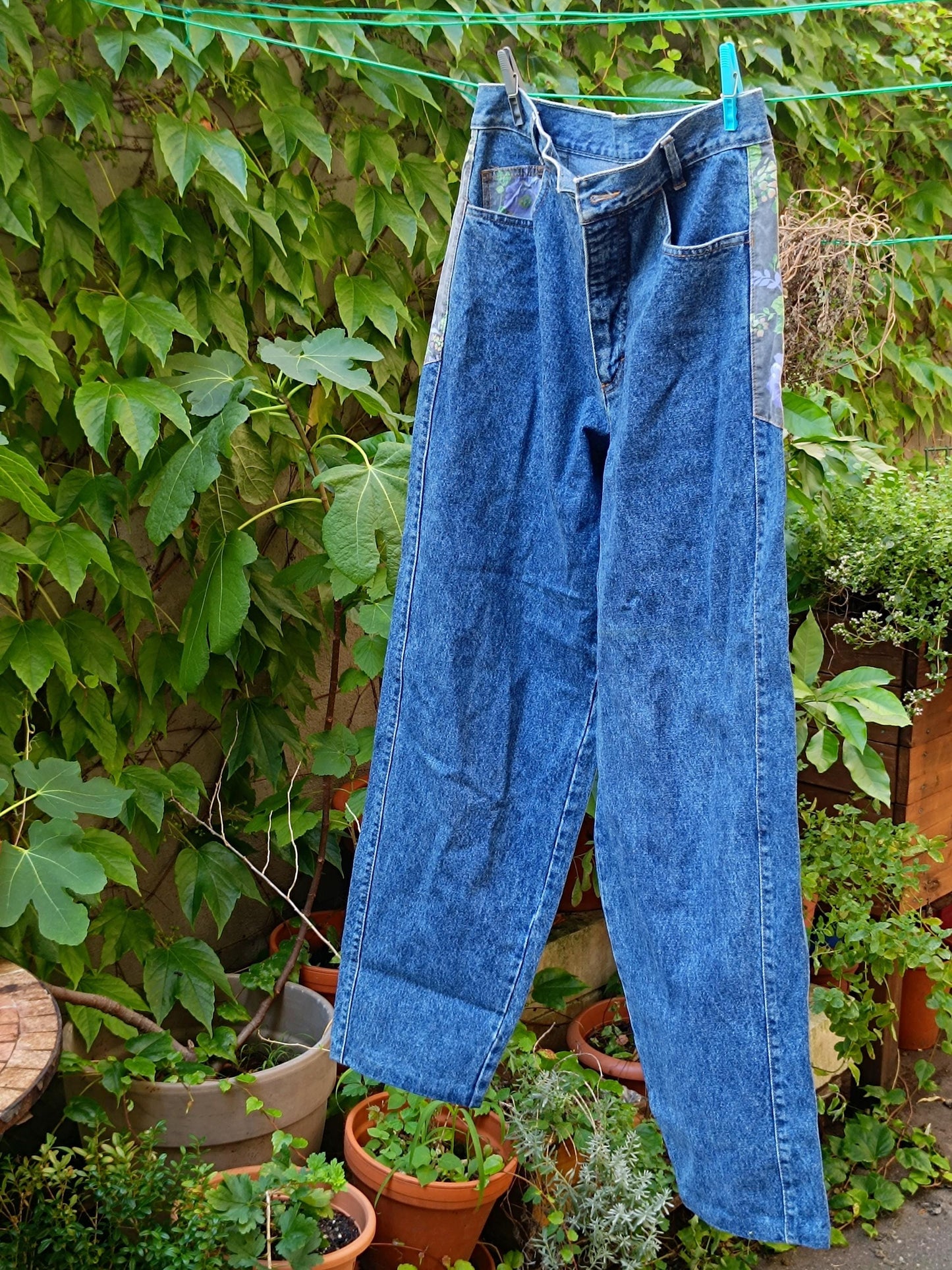 A still life image of a pair of vintage high-waisted blue jeans by the brand vicini. The blue jeans from the 80s hang on a laundry line with two clothing pegs against a leafy background.