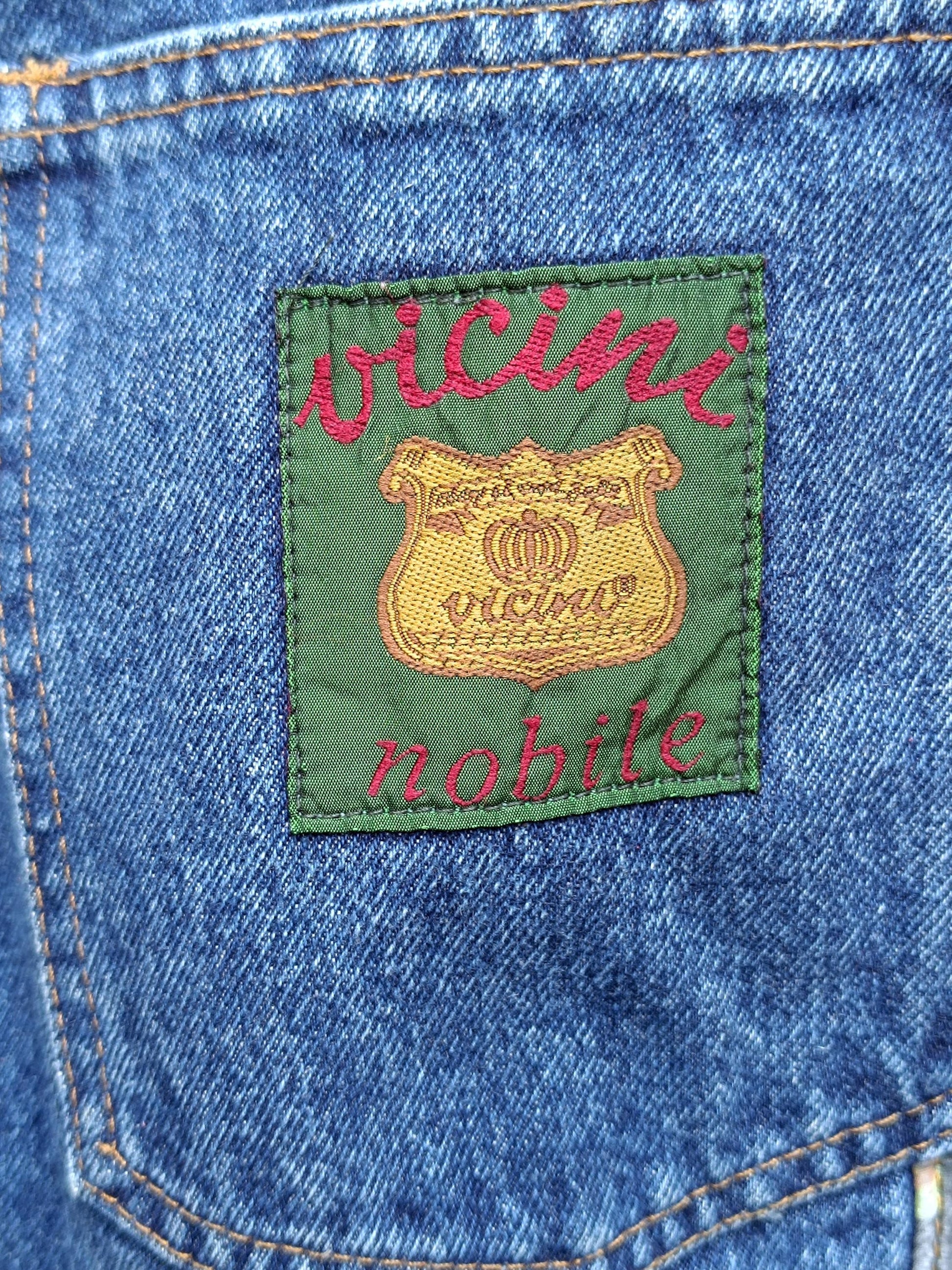 A close up still life image of the logo of a pair of vintage high-waisted blue jeans. The green with red leathering logo reads: vicini.