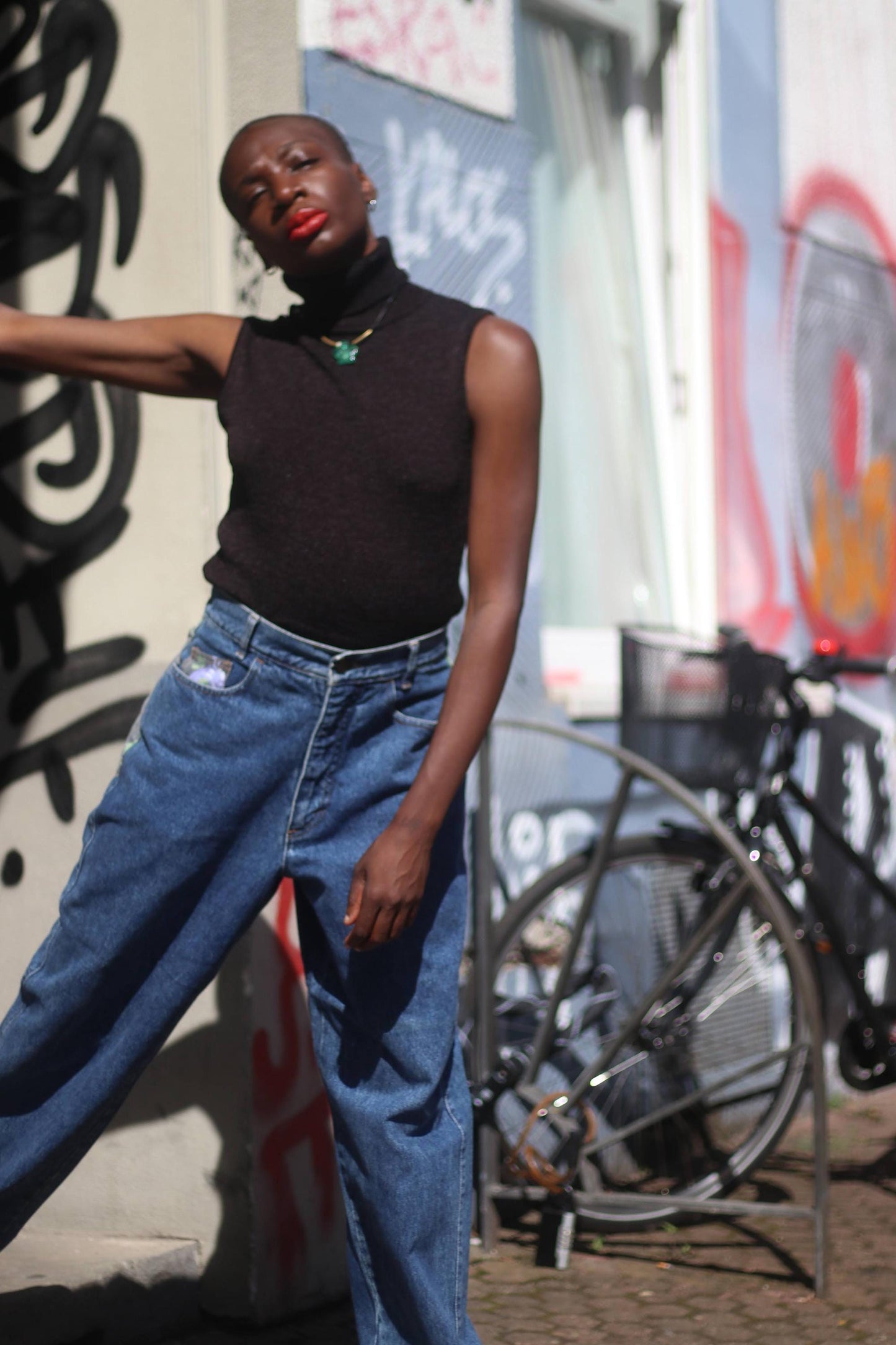 A black female model in vintage high-waisted jeans from the 80s and a black sleeveless top photographed with a graffiti in the background. The jeans have floral patchwork side details