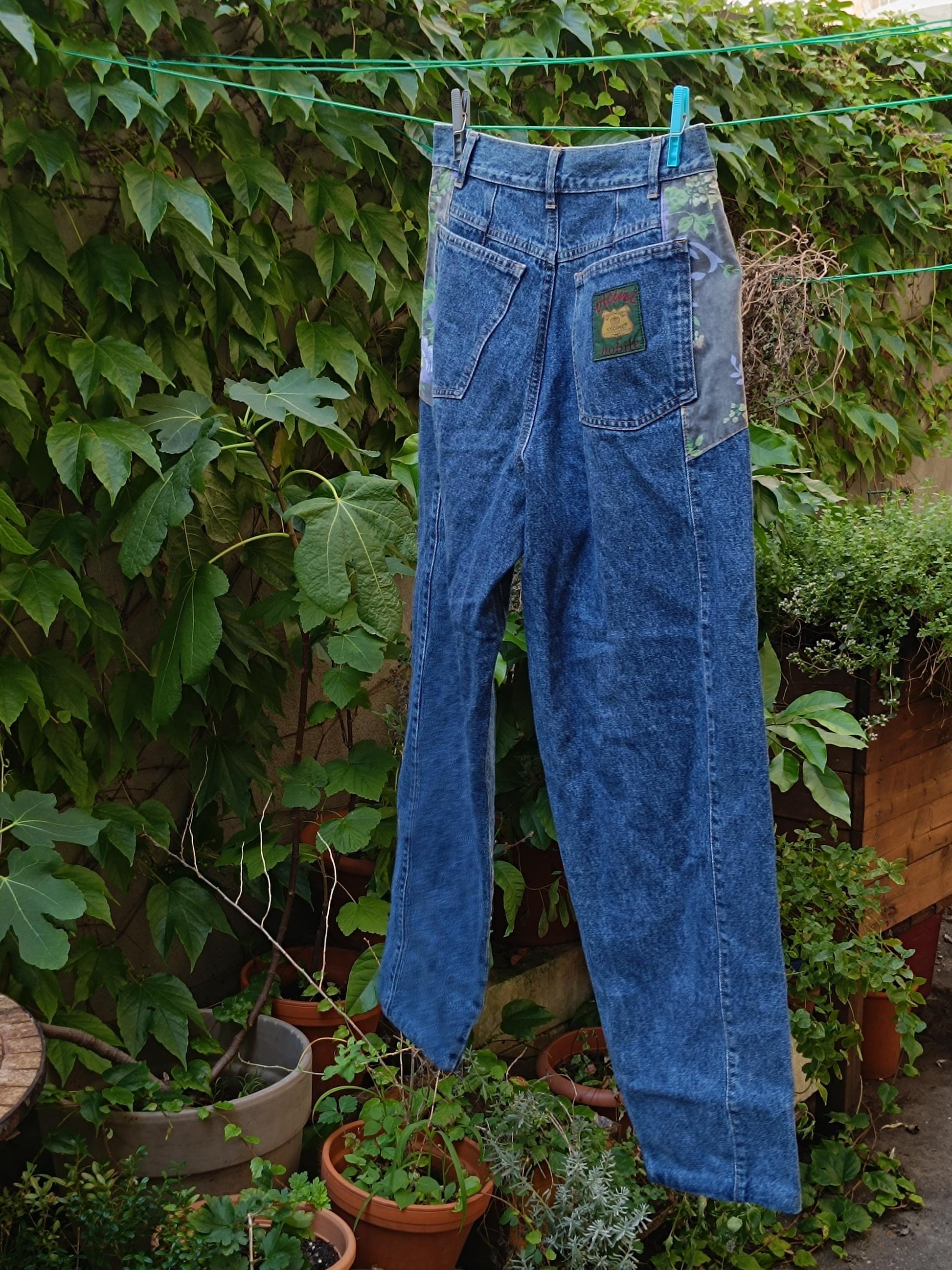 A still life image of a pair of vintage high-waisted blue jeans by the brand vicini. The blue jeans from the 80s hang on a laundry line with two clothing pegs against a leafy background.