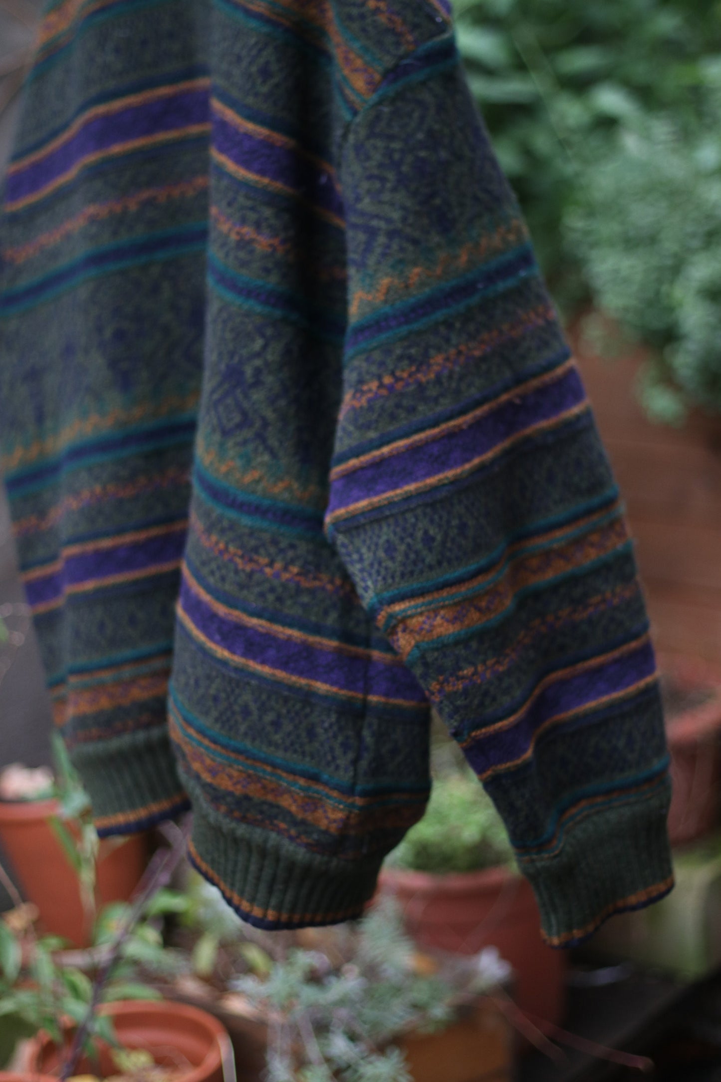 Back view of cozy multi-colored cardigan with green, orange, and purple stripes hanging on a hanger outdoors against a concrete wall with climbing vines. The long-sleeved vintage design can be worn as a coat it is perfect for fall and winter fashion