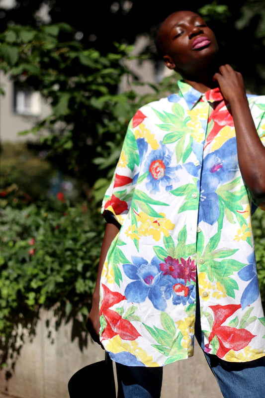 A female model of African heritage poses pensively  in a colorful floral print shirt and jeans. She is photographed against a green leafy background, touching the collar of the buttoned down shirt and  her eyes are closed