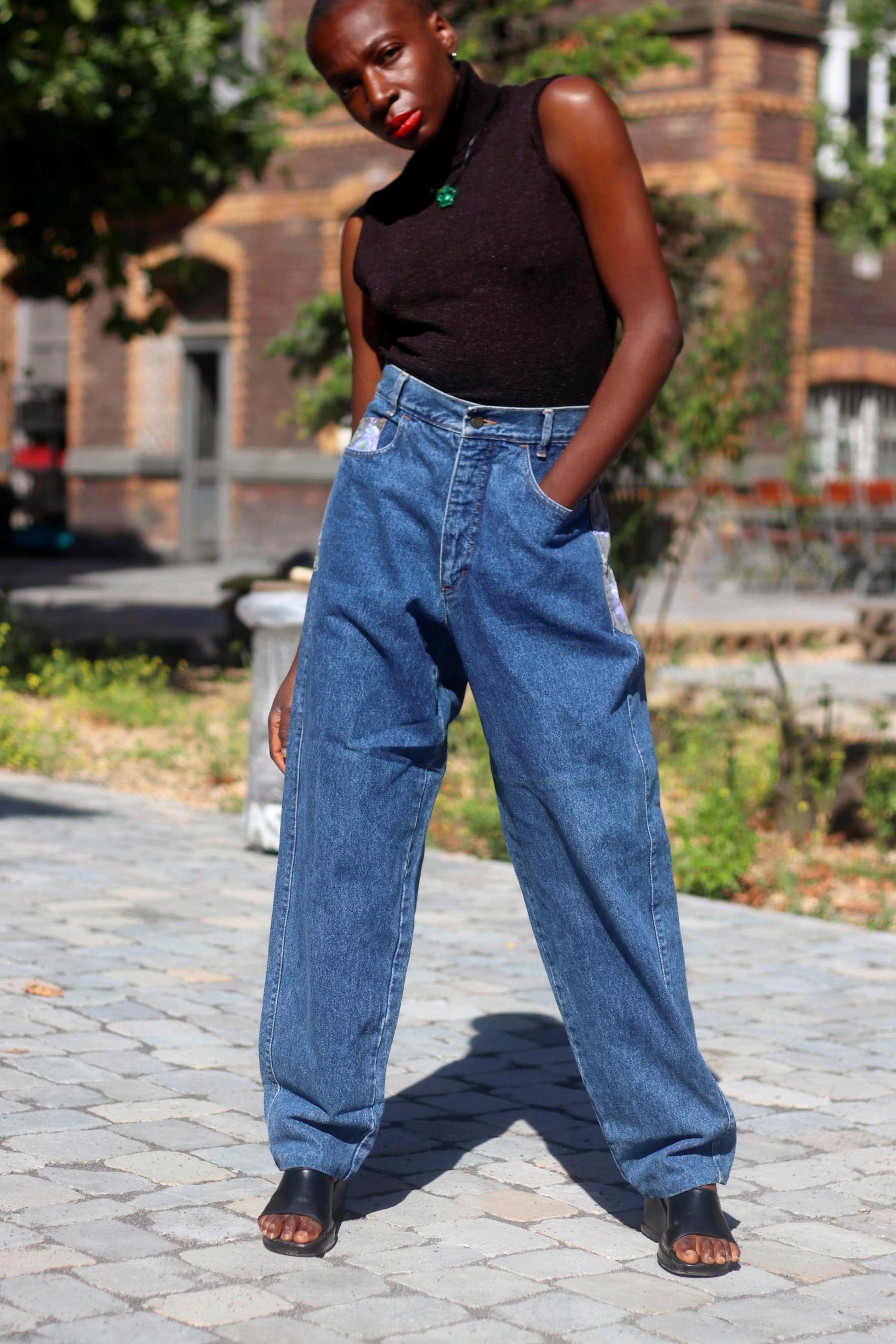 A black woman in a pair of vintage high-waisted jeans from the 80s and a black sleeveless top stands confidently against an urban background showcasing a stylish outfit. She has one hand in her pocket and the jeans have floral patchwork side details