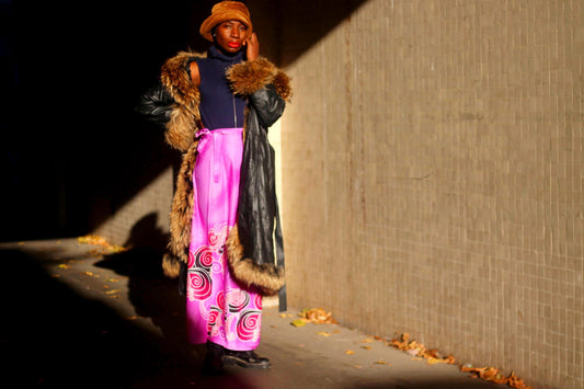 Female in a long black leather coat with fur trim, a brown hat, and a vibrant vintage pink skirt featuring swirling patterns. They wear a dark turtleneck, and red lipstick, posing confidently in a dimly lit urban setting.