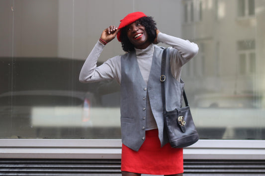 Model of African heritage wearing a red linen skirt styled with a grey turtleneck and vest, accessorized with a red beret and black leather back. She is photographed against a glass window with a whimsical smile.