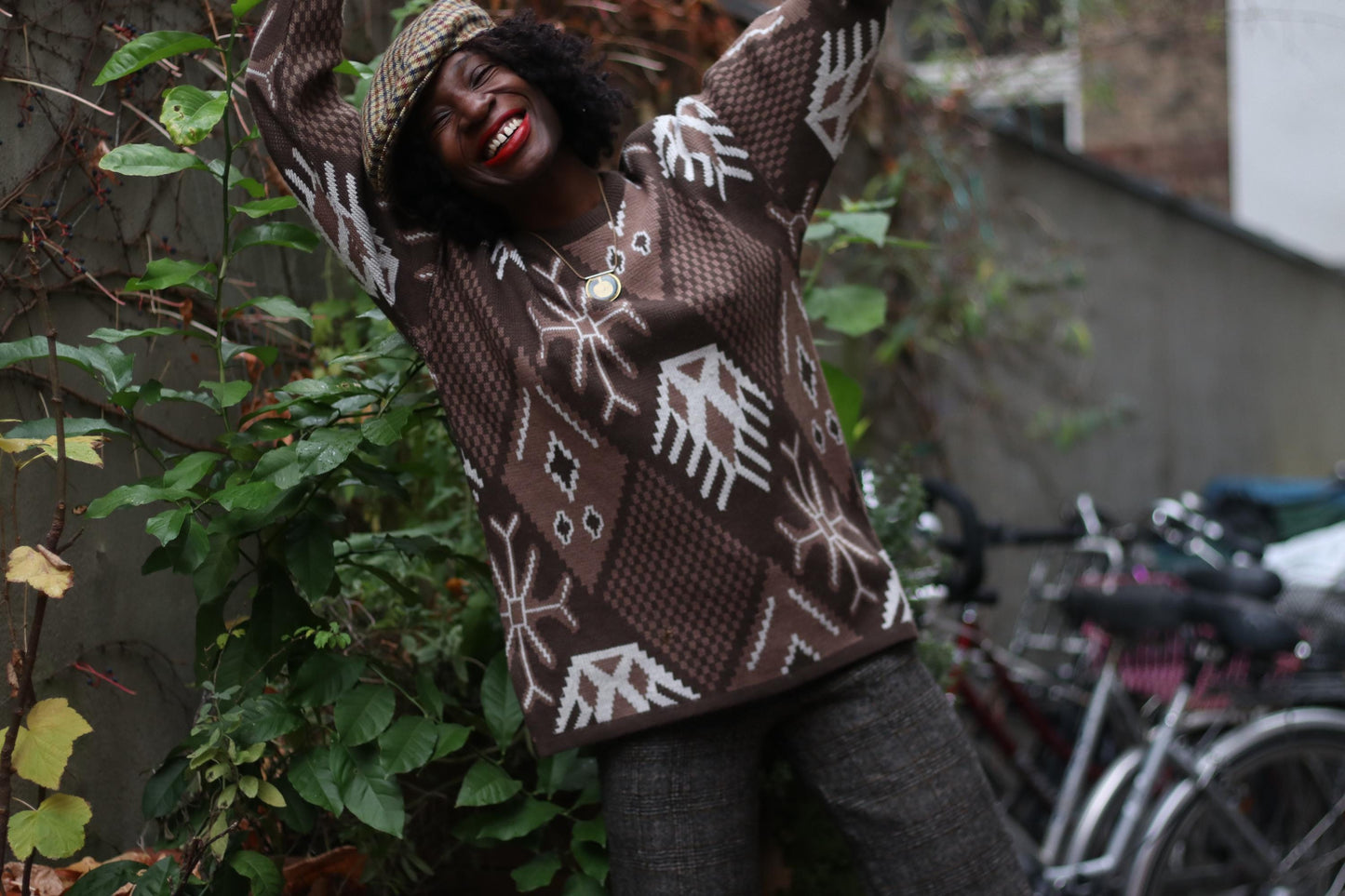 A person smiles brightly with arms raised, wearing an 80s Ciro Esposito sweater—a vintage, quirky Christmas jumper with white snowflake patterns. The long-sleeved, hipster-style sweater stands out against a backdrop of green foliage and bicycles.