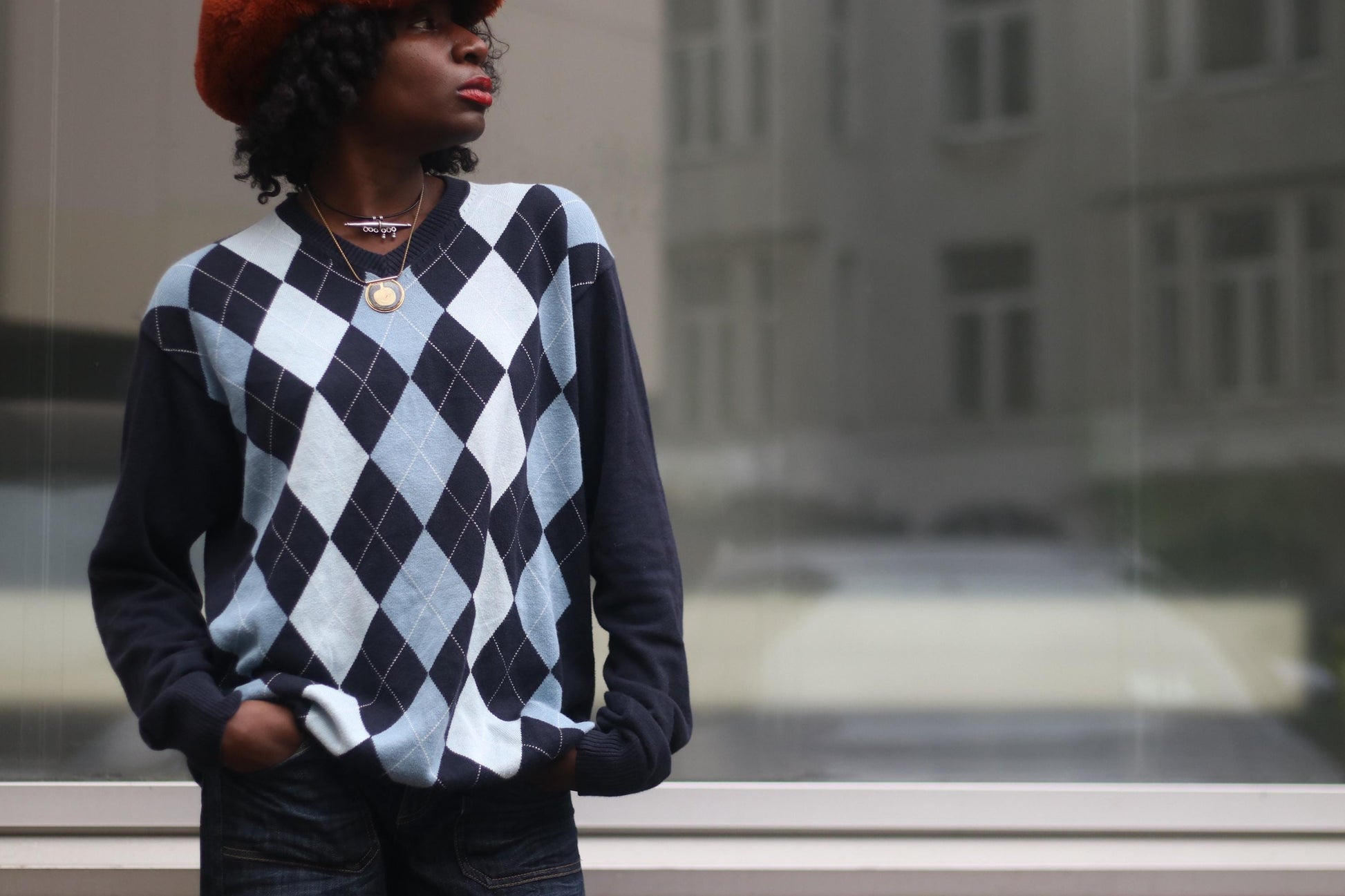 Model wearing a vintage 90s blue argyle print sweater, standing outdoors against a neutral glass window. The sweater features a classic argyle pattern in blue, gray, and white, with a relaxed fit and ribbed cuffs and hem. The model  pairs the sweater with high-waisted jeans for a casual, retro-inspired look.