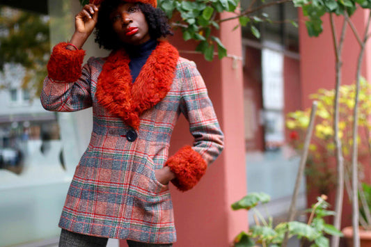 A black model stands outdoors near a pink building, wearing a vintage Gil Bret plaid coat with burnt-orange faux fur collar and cuffs. Green plants are visible nearby, .The coat has a single button and front pockets