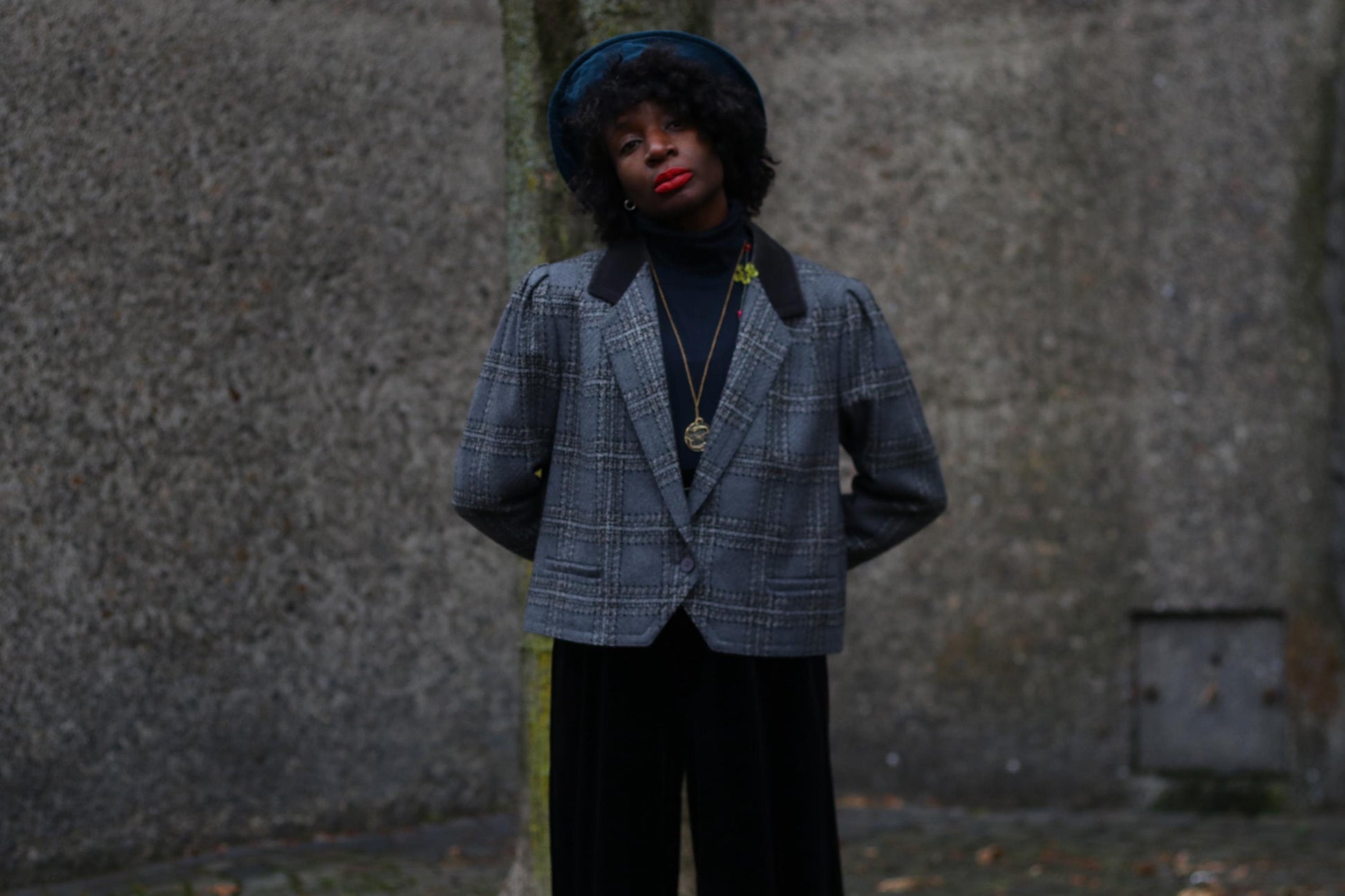 Model of African heritage wearing a vintage stylish grey plaid blazer and blue hat, standing against a textured stone wall, showcasing vintage-inspired street style. The wool blazer is by the german brand Miss Antoinette Franz Haushofer