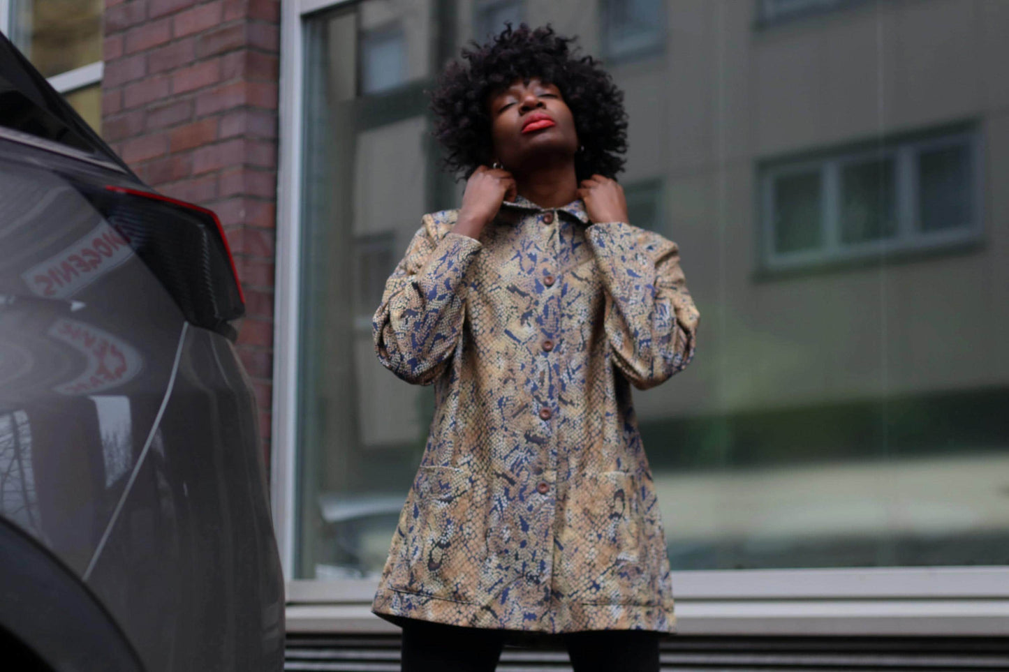 Fashionable individual in a stylish snake print jacket, standing outdoors next to a modern car. The person is posing thoughtfully with one hand touching the collar of the jacket. Urban street style fashion concept.