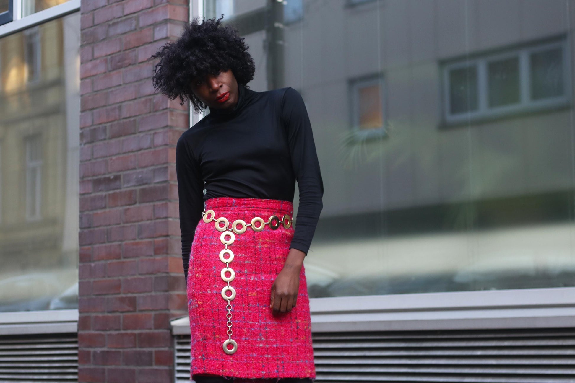 Fashionable black woman wearing a black turtleneck and a 90s Pink Tweed Skirt with a gold chain belt, posing against an urban city backdrop.