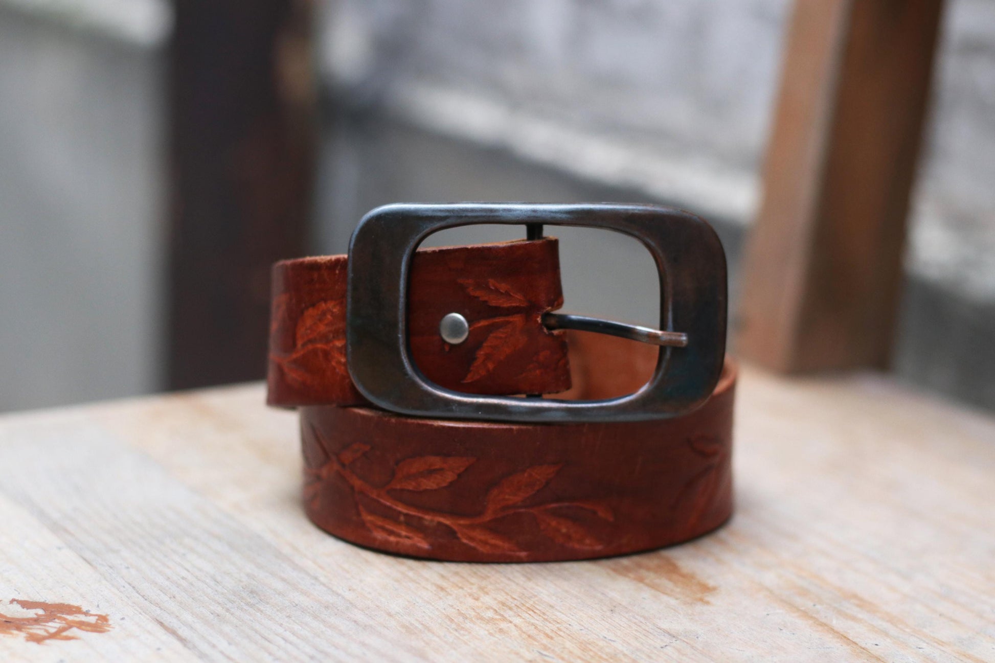 Vintage brown leather belt with an engraved leaf pattern and a rustic metal buckle on a wooden surface.