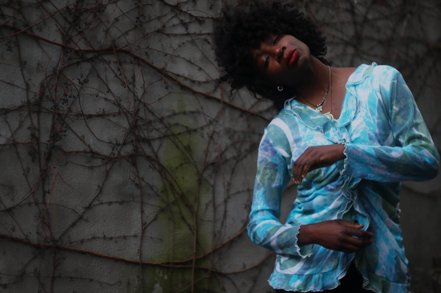 A Fashionable model of African descent  in a vintage y2k blue patterned blouse posing against a textured, vine-covered wall.