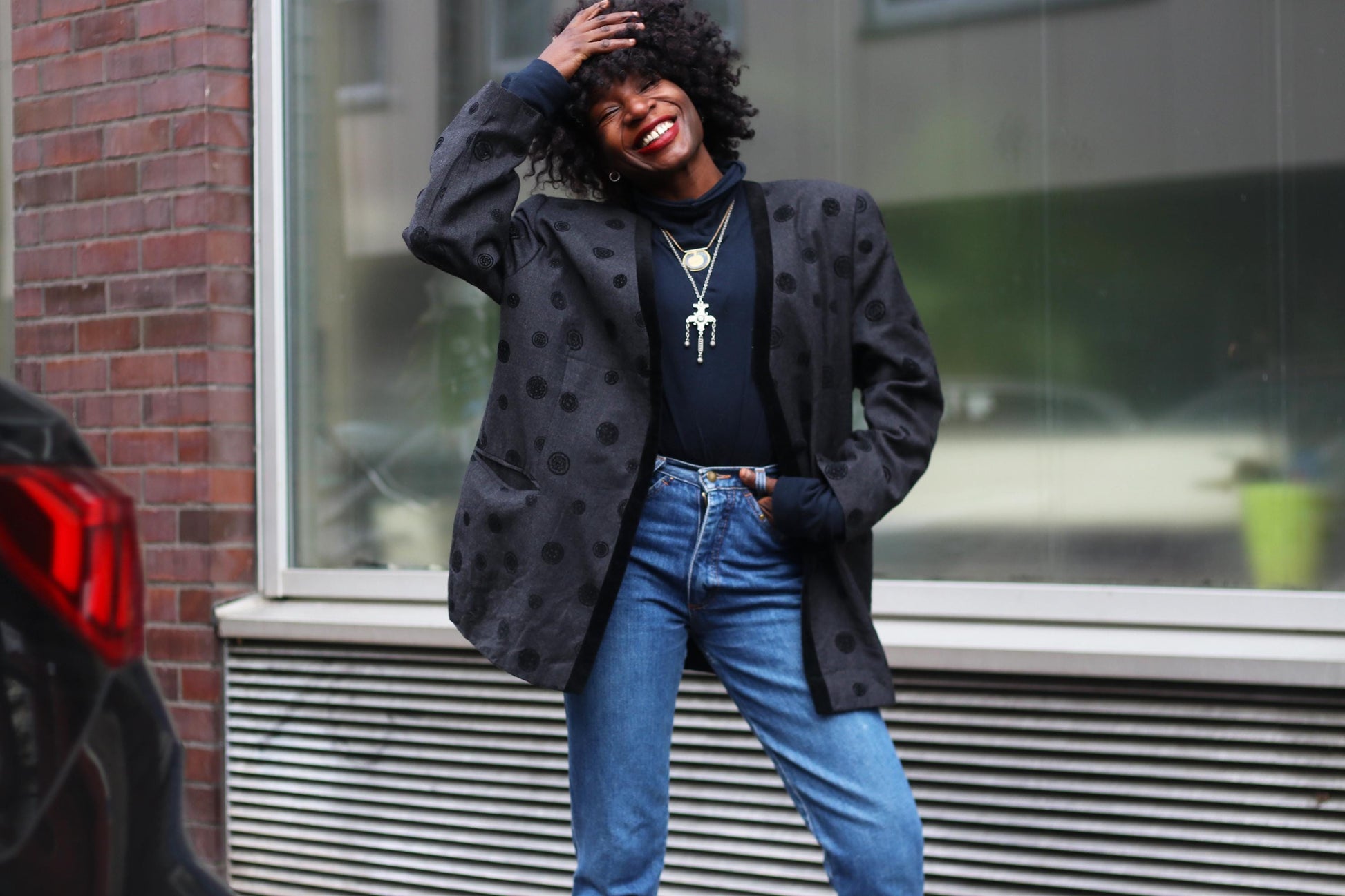 Fashionable woman in a vintage 90s polka dot grey blazer paired with high-waisted blue jeans poses confidently in an urban setting, showcasing casual street style with layered necklaces and a joyful smile