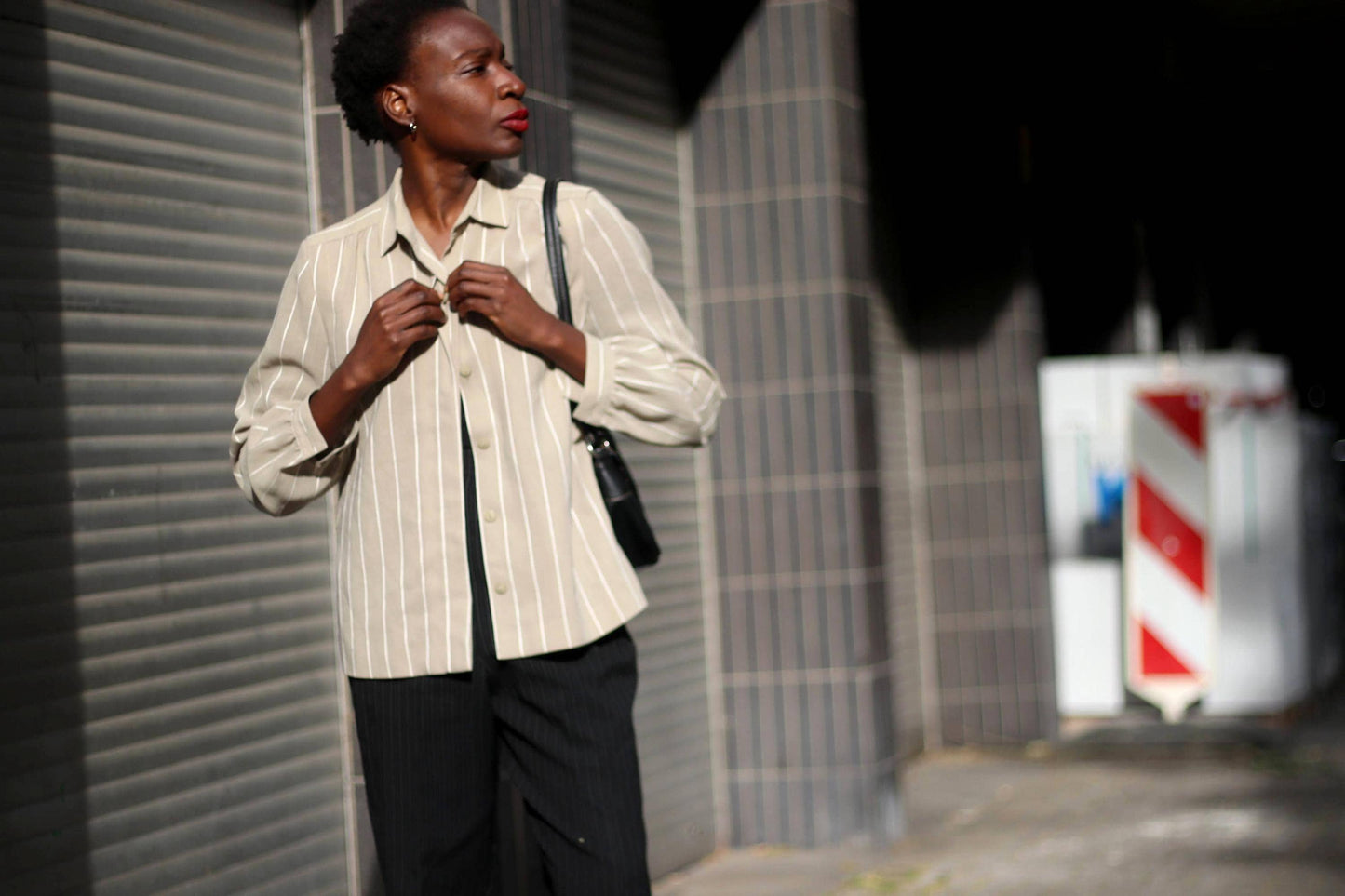 Fashionable woman with a small afro wearing a vintage striped grey styled white blouse with pinstriped black trousers.She is photographed against a grey urban wall with blocked pattern. The imagery oozes a streetstyle vibe.