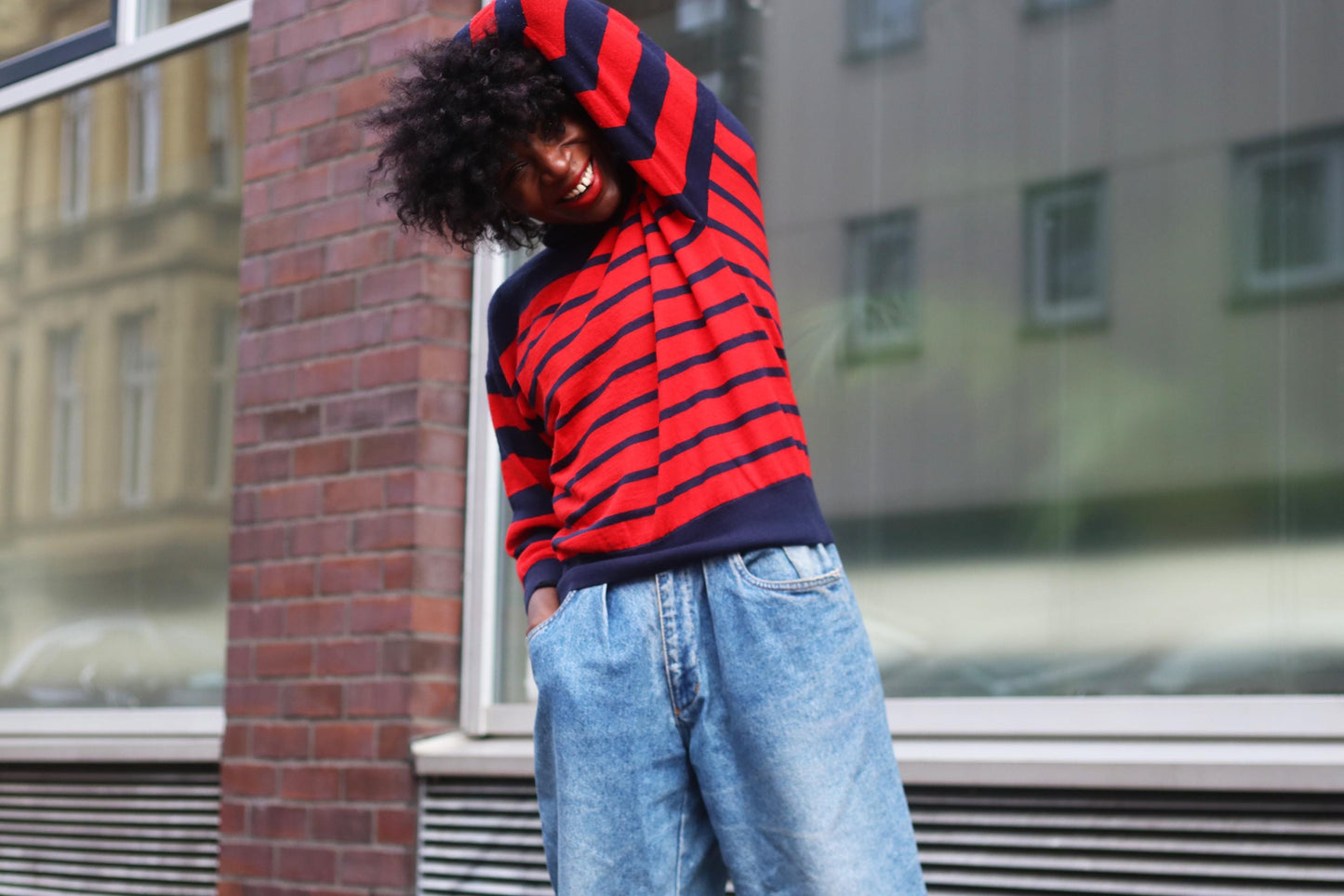 Fashionable female  smiling in urban setting, wearing a vintage 90s Roberto Sarto stylish red and navy striped sweater with loose-fitting denim jeans. Casual fashion streetwear photography.