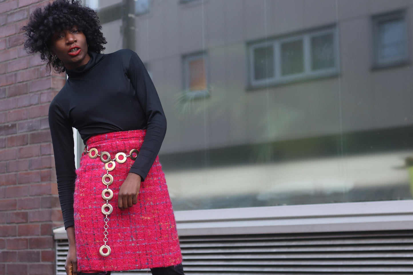 Women wearing a pink tweed mini skirt and a black turtleneck. The skirt is accessories with a gold-toned chain link belt. She is photographed outside standing in front of a big window.