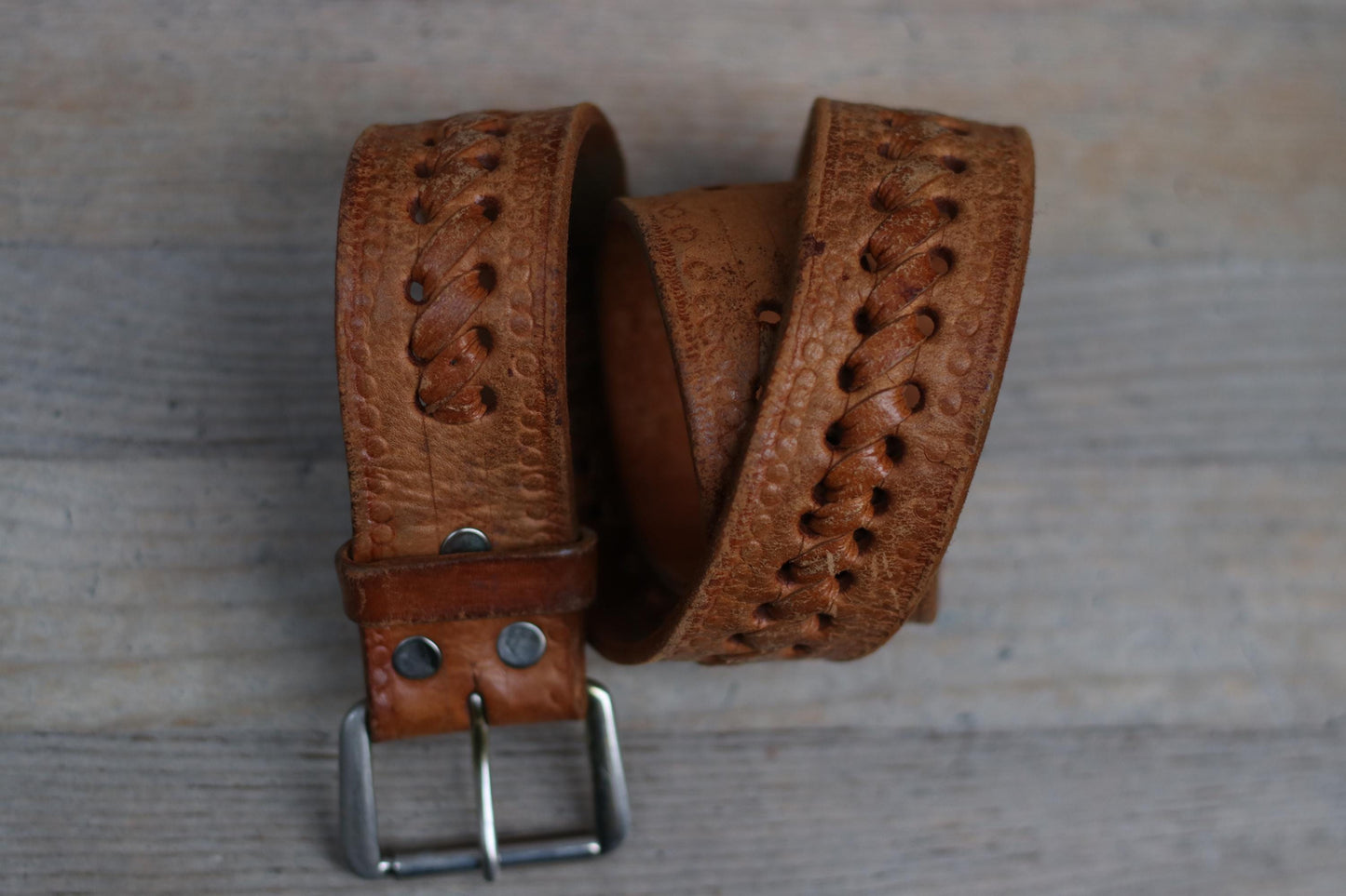 A vintage brown Western woven leather belt, showcasing its intricate handwoven pattern, with a classic cowboy-style silver buckle is displayed on a rustic wooden surface. The belt&#39;s rugged charm and authentic Western style are highlighted.