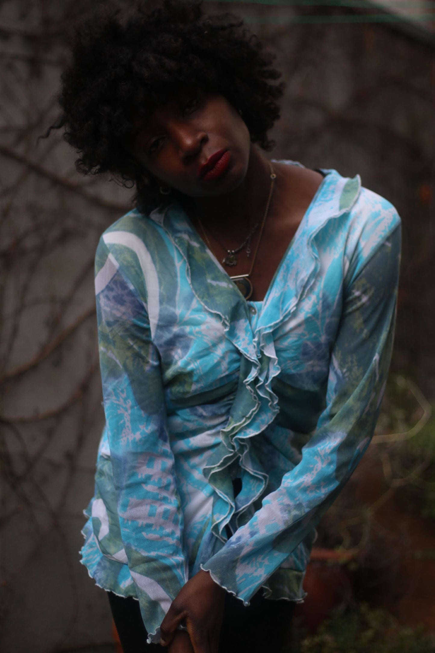 Portrait of a black female model wearing a vintage y2k  patterned turquoise blouse, with a thoughtful expression, standing outdoors against a backdrop of vines.