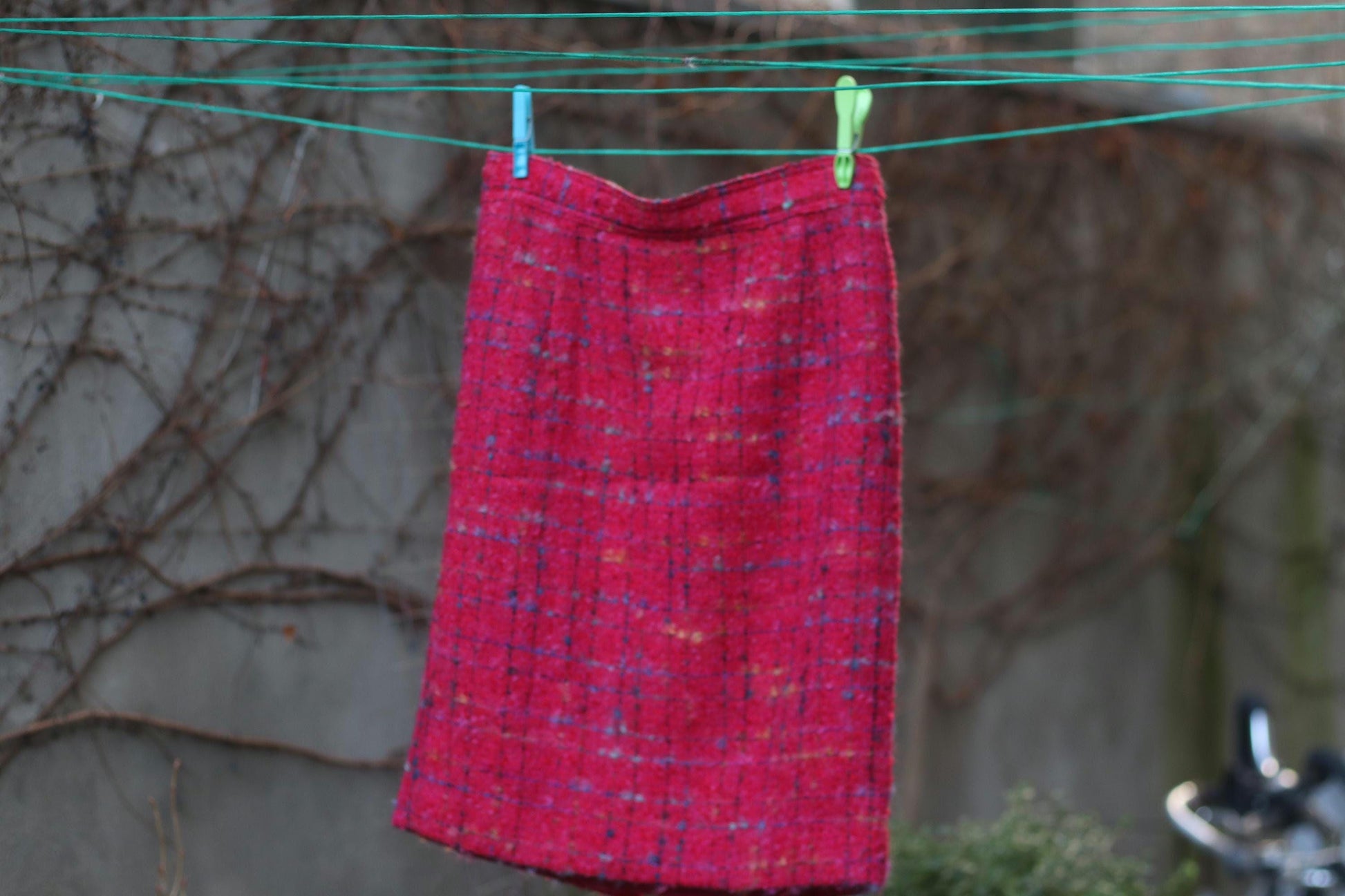 A still live imagery of a  vintage pink mini skirt from the 90s. The  preppy chic design is photographed on a laundry line held up by two clothes peg against a background with brown vines.