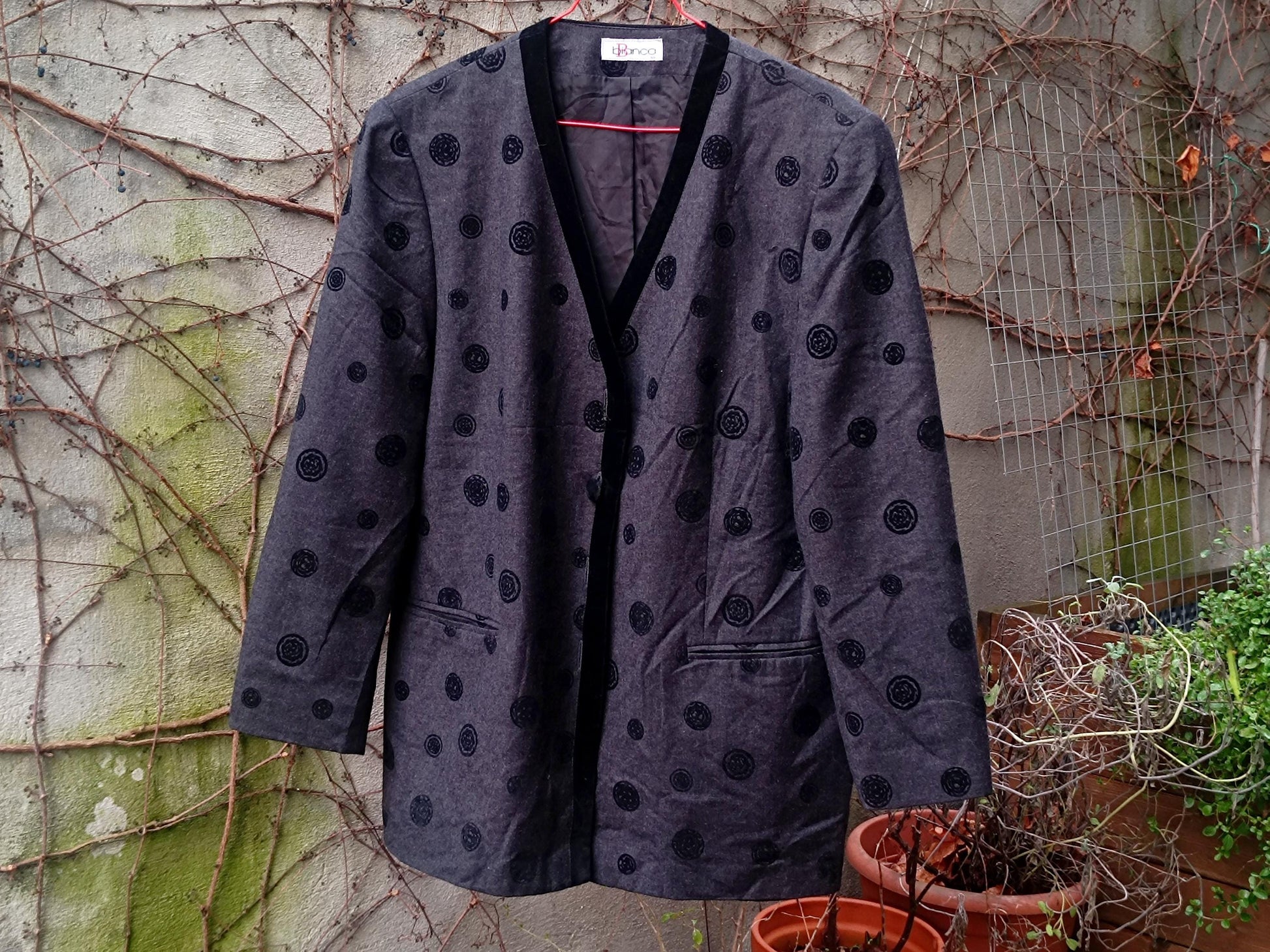 Close-up of a gray and black polka-dotted blazer showing the texture of the fabric and the details of the pattern, with a blurred background of climbing vines and potted plants.