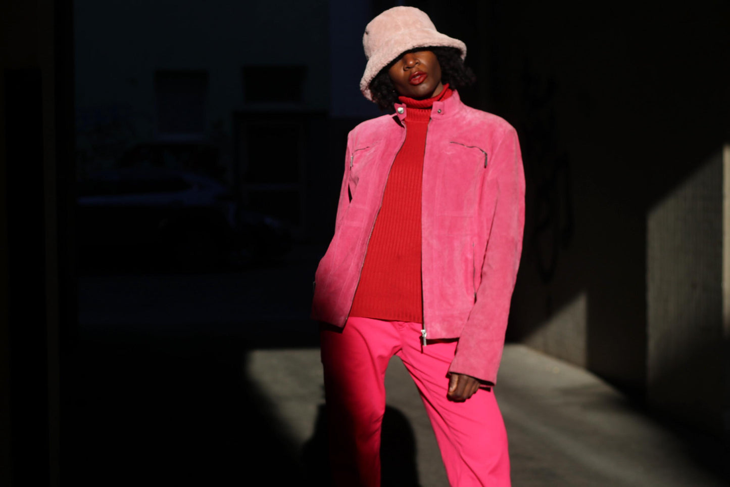 A woman in a vintage pink suede jacket by Alma, paired with a red turtleneck and pink trousers and textured bucket hat, posing confidently in moody lighting for a high-fashion street style look.