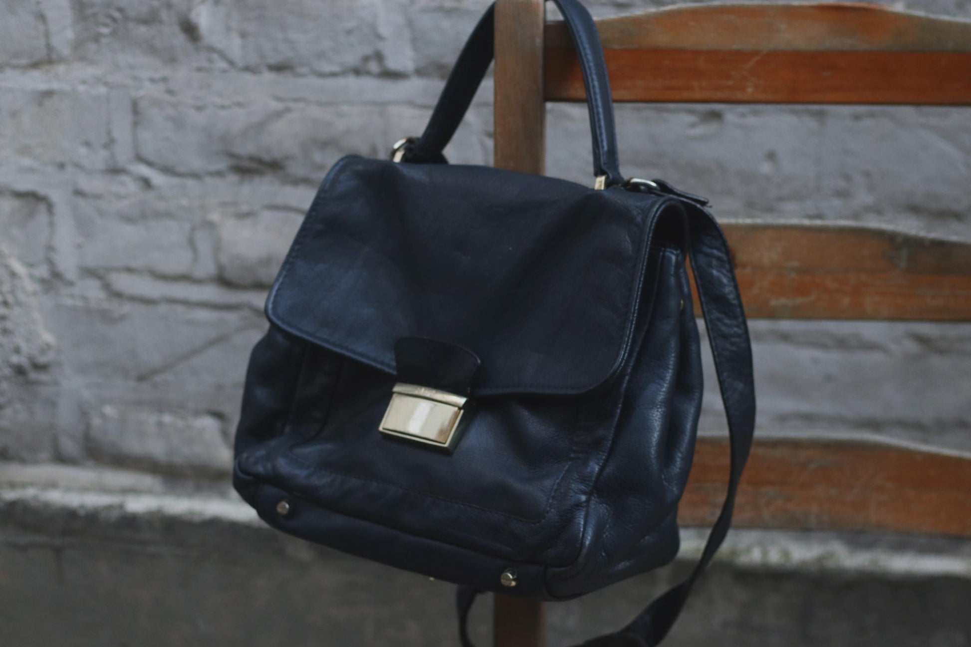 90s vintage black leather shoulder/crossbody bag with a top handle hanging  from a wooden chair. The  supple leather, classic flap shape, top handle, and crossbody strap make it a versatile accessory. A grey textured wall provides a simple backdrop.