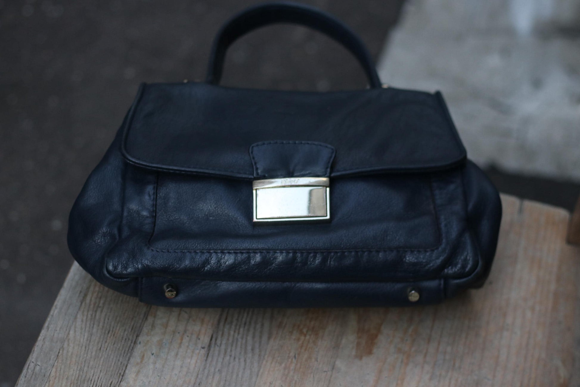 90s Black leather top-handle bag  by Abro with silver-toned hardware on a wooden surface. Classic design featuring a flap closure and visible stitching. Soft, worn texture suggests vintage quality. Focus on the details and craftsmanship.