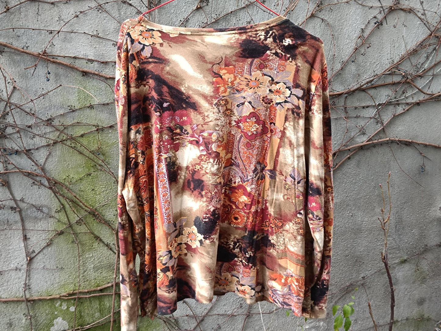 Vintage floral blouse in taupe, rust, and peach tones. Abstract floral print, boho style, round-neck, and lightweight. it&#39;s on a hanger against a textured grey and green background.